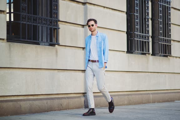 Man in light blue shirt and sunglasses walking past a stone building with iron window grilles.