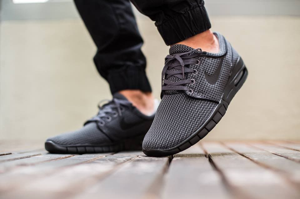 Nike Janoski Max sneakers in dark grey and black worn by a person on a wooden deck.