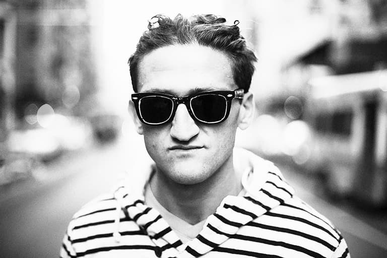 Casey Neistat wearing sunglasses and a striped hoodie, standing on a city street with a blurred background.