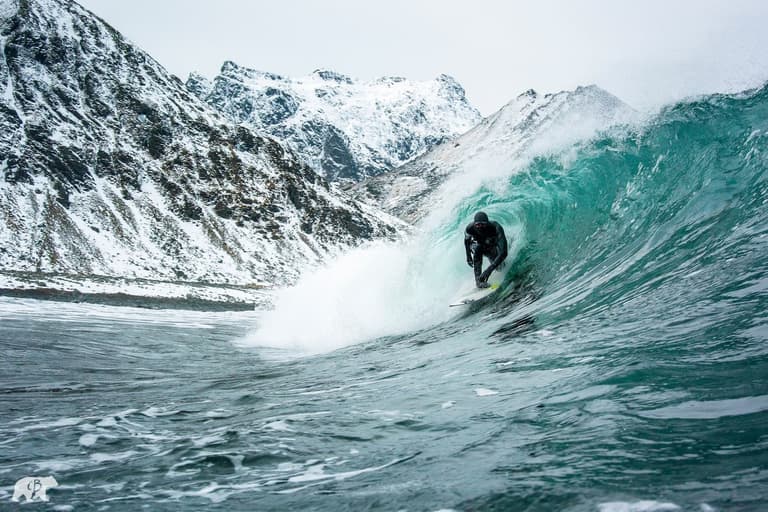 Surfer rides a turquoise wave near snow-covered mountains under a cloudy sky.