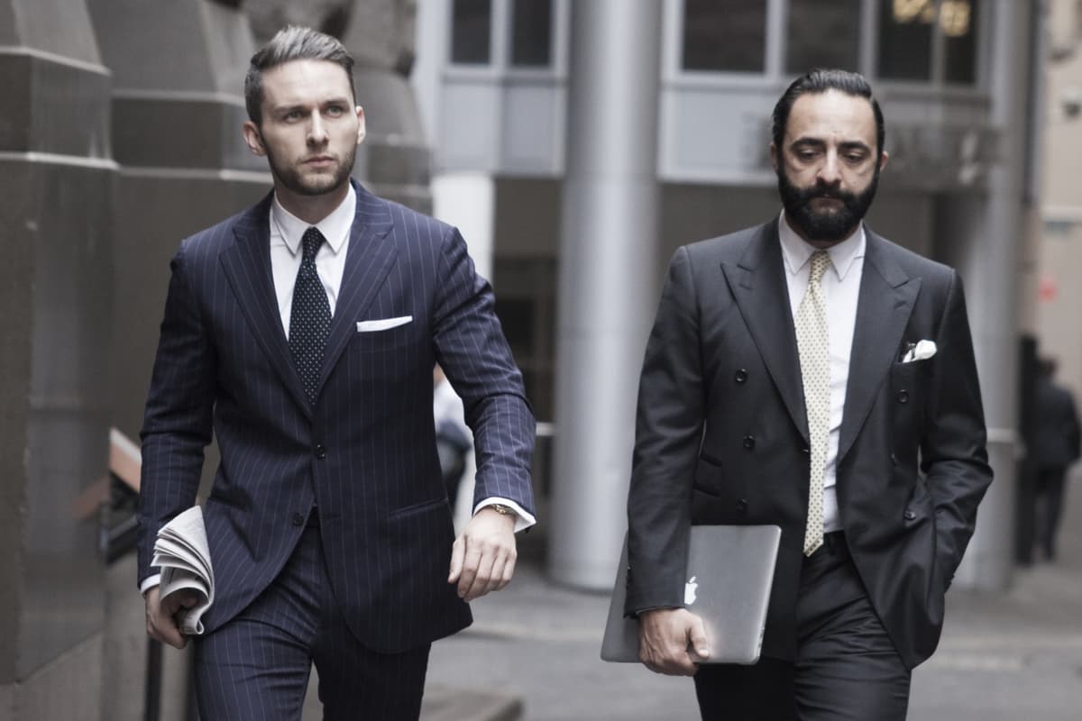 Two men in tailored suits walking in an urban setting, one holding a newspaper, the other a laptop.