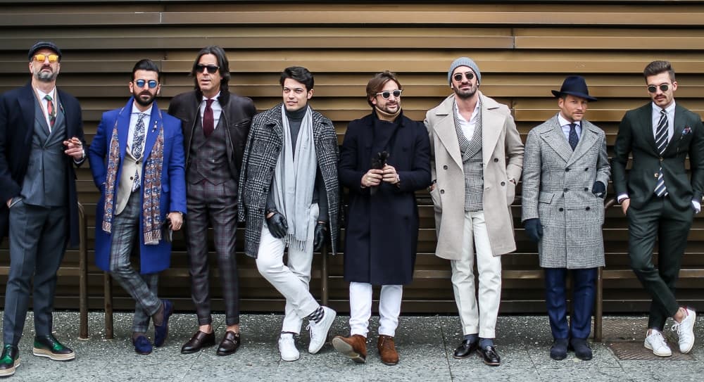 Group of stylish men in diverse suits and coats, posing against a modern brown wall at Pitti Uomo 91.