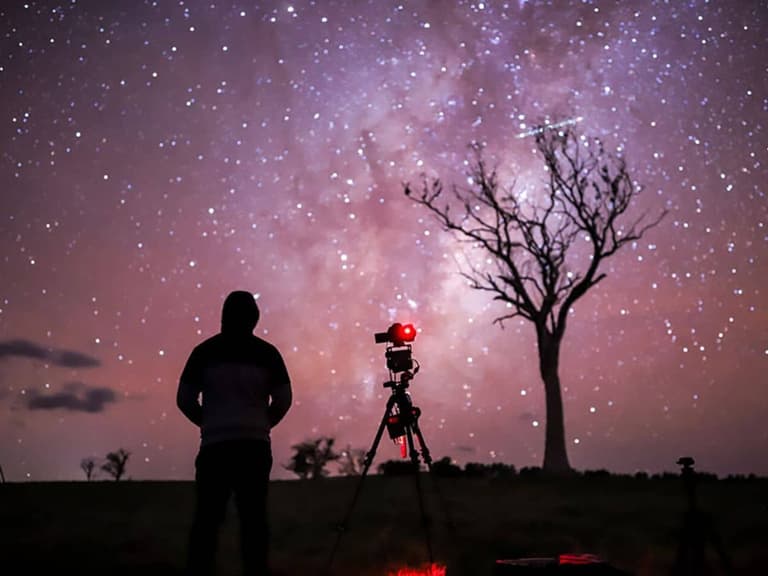 Silhouetted photographer with Canon camera capturing starry night sky and tree under purple hues.