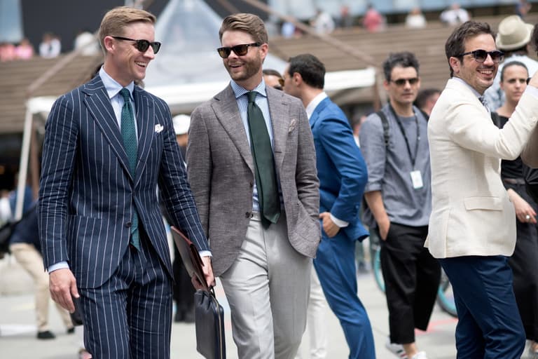 Men in stylish summer suits walking outdoors, wearing sunglasses and smiling, with a crowd in the background.