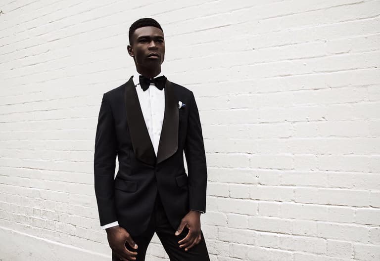 Man in black tuxedo with satin lapels stands against a white brick wall, wearing a bow tie and pocket square.