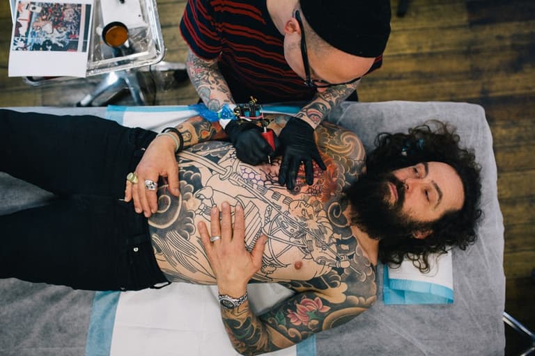 Tattoo artist in striped shirt working on man's chest tattoo in studio with wooden floor.