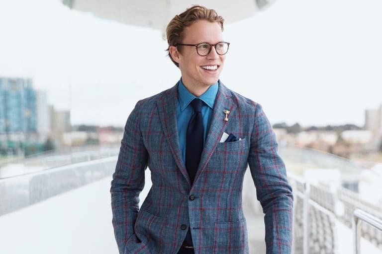 Dalton Henshaw wearing a blue plaid suit and glasses, smiling on a balcony with a cityscape background.