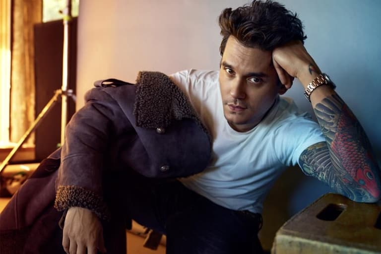 John Mayer wearing a white T-shirt and dark jacket, seated with a thoughtful expression in a studio setting.