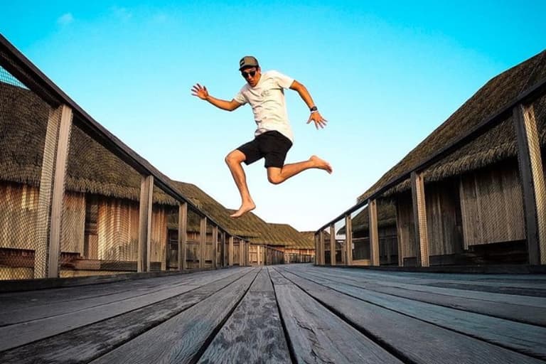 Person jumping on a wooden walkway between thatched-roof huts under a clear blue sky.