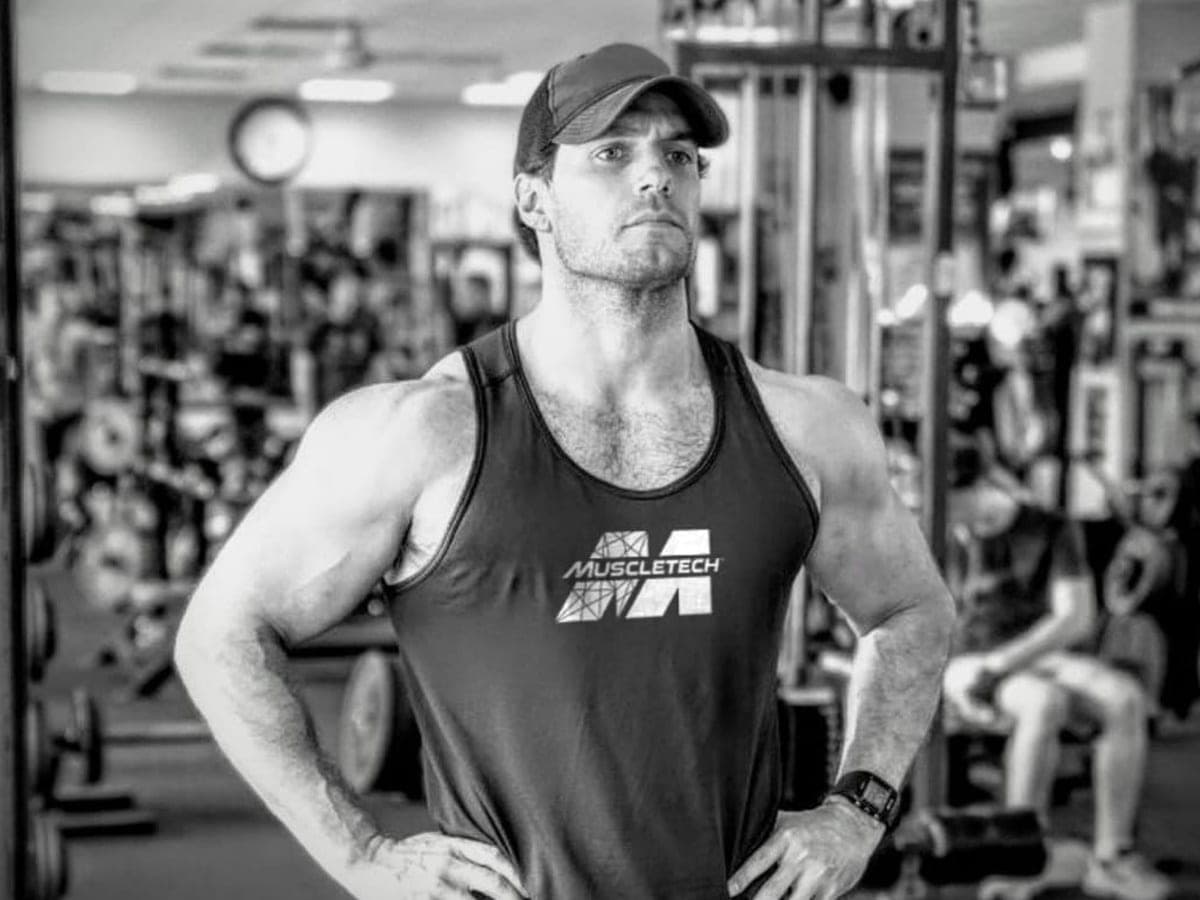 Henry Cavill wearing a Muscletech tank top and cap, standing confidently in a gym with hands on hips.
