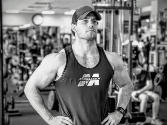 Henry Cavill wearing a Muscletech tank top and cap, standing confidently in a gym with hands on hips.
