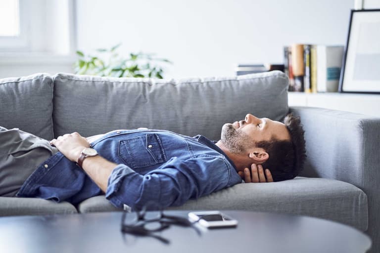 Man in denim shirt sleeping on grey sofa with smartphone on table in bright living room.