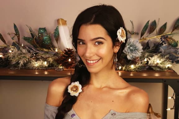Asmr artist smiling with floral hair accessories, seated in front of decorative holiday foliage and warm lighting.