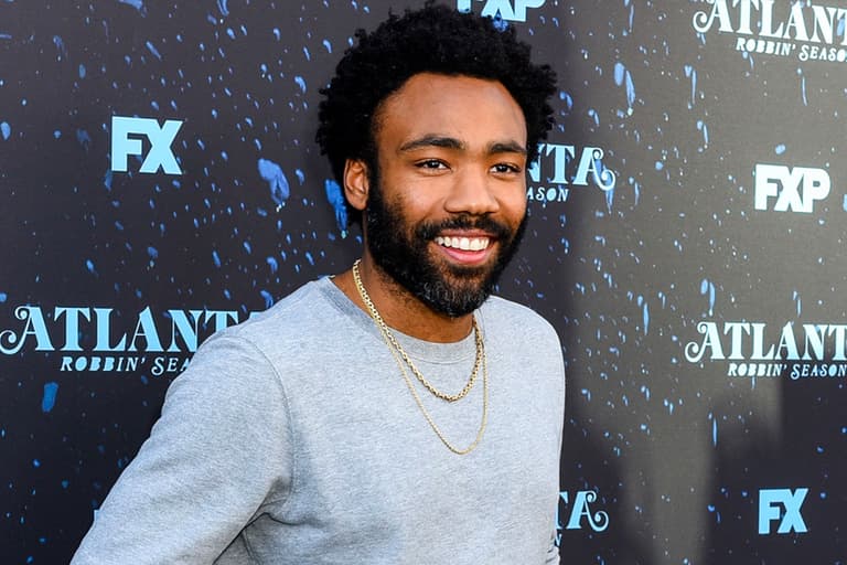 Donald Glover smiling in a grey t-shirt with gold chains at an Atlanta event backdrop.