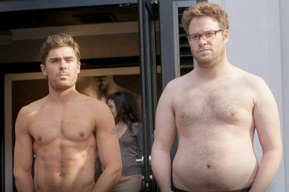 Two shirtless men standing outside, one with defined abs and the other with a softer physique.