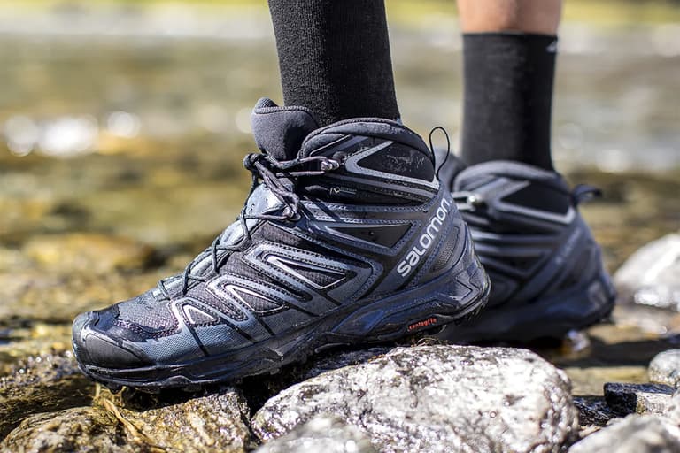 Salomon hiking boots in black and blue worn by a person standing on wet rocks near a stream.