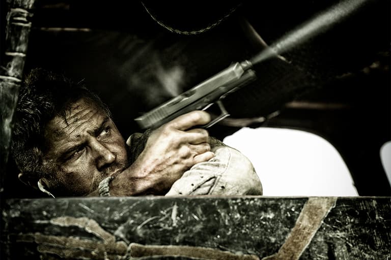 Tom Hardy aiming a gun from a vehicle in Mad Max: Fury Road, with intense expression and dusty setting.