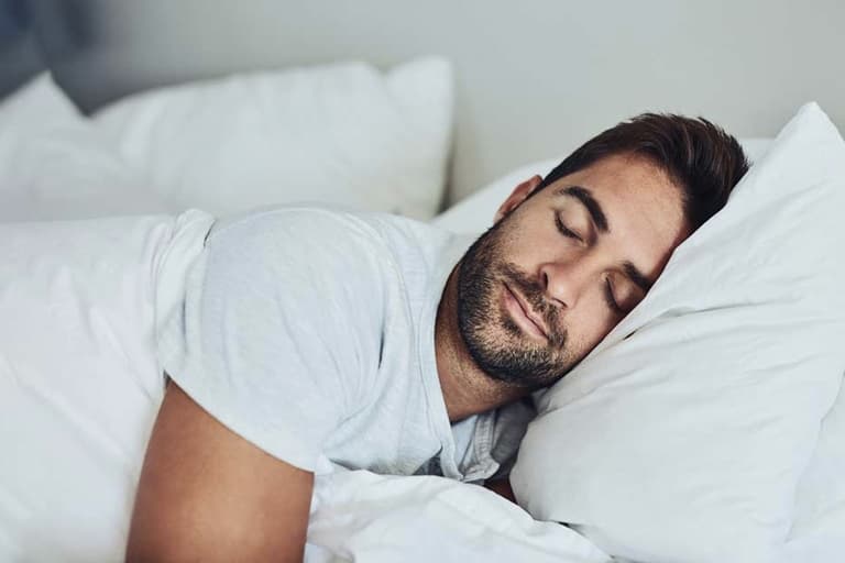 Man sleeping peacefully on white pillows and sheets in a softly lit bedroom.