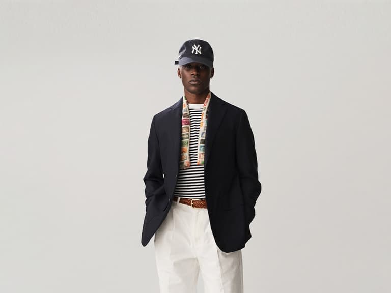 Man wearing a dark blazer, striped shirt, and white trousers with a cap, standing against a plain background.