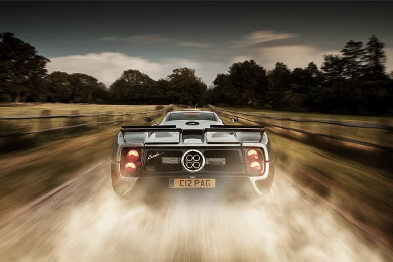 Pagani Zonda rear view on country road with motion blur and dust trail, surrounded by trees.