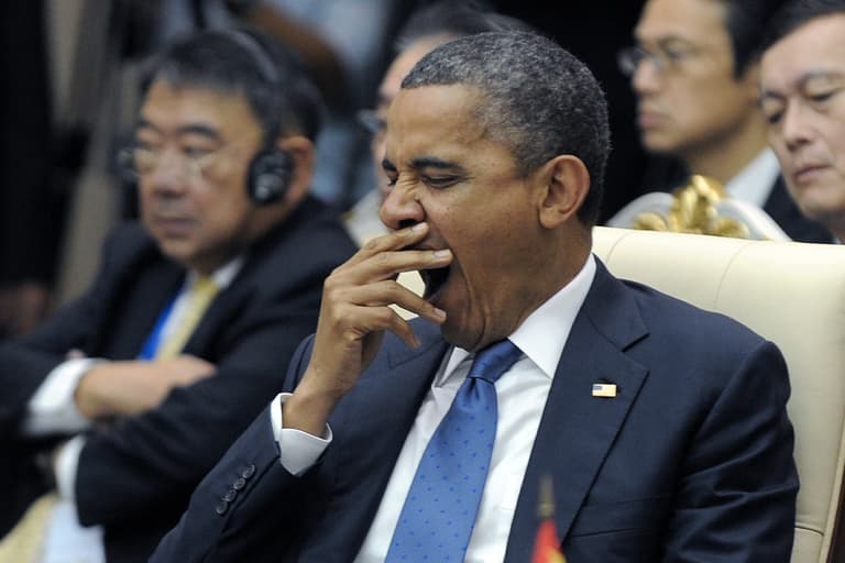 Man in a suit yawning during a conference, seated next to a person wearing headphones.