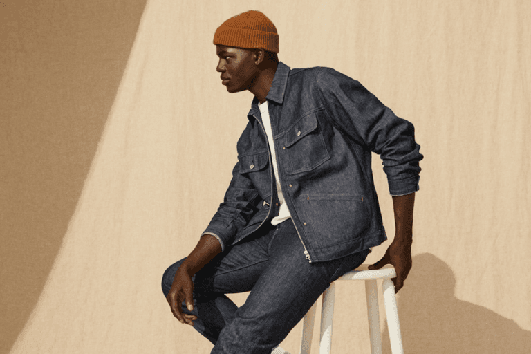 Model wearing H&M denim jacket and jeans with a white t-shirt, sitting on a stool against a beige backdrop.
