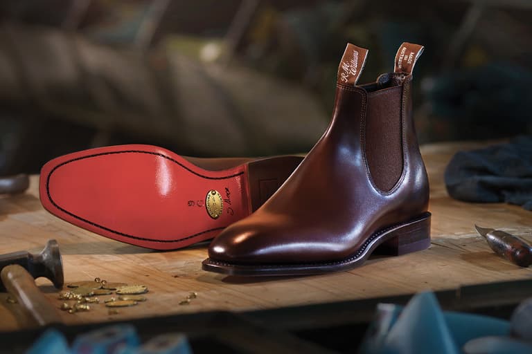 RM Williams brown leather boots on a wooden workbench, showcasing red sole and signature pull tabs.