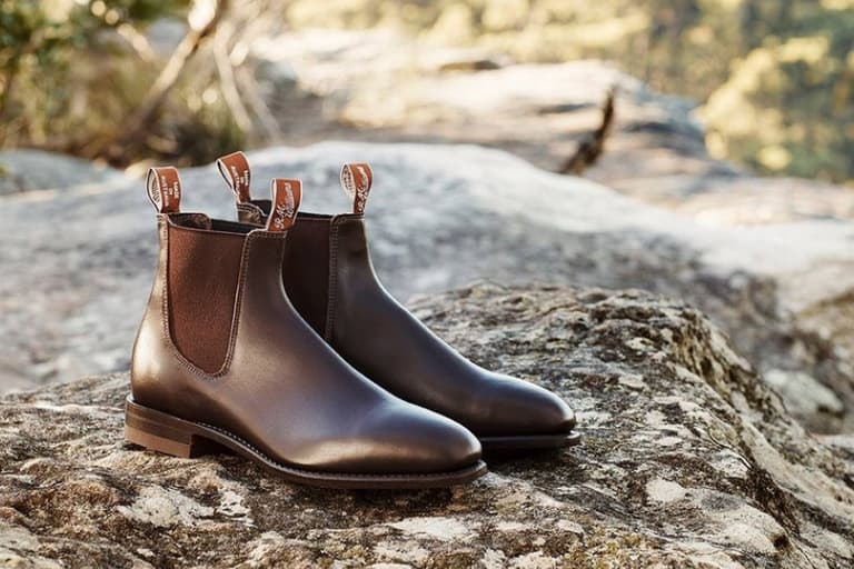 R.M. Williams brown leather boots resting on a sunlit rock in an outdoor setting.