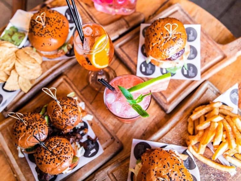 Burgers and fries on wooden boards with cocktails, featuring sesame seed buns and vibrant garnishes.