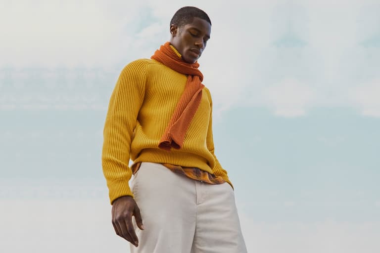 Man wearing yellow sweater and orange scarf against a cloudy sky, looking down with hands in pockets.