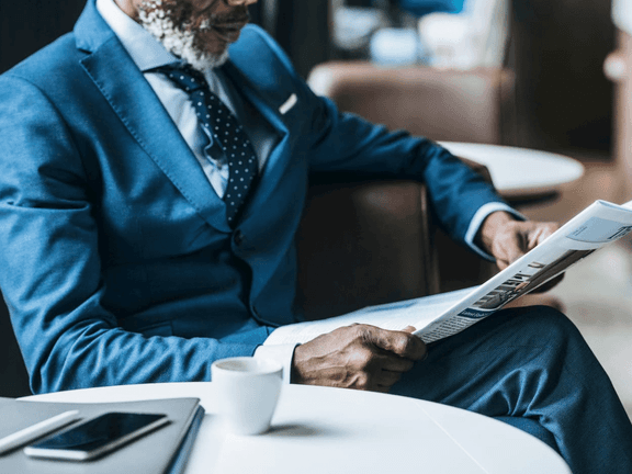 Man in blue suit reading newspaper at a cafe table with a coffee cup and laptop nearby.