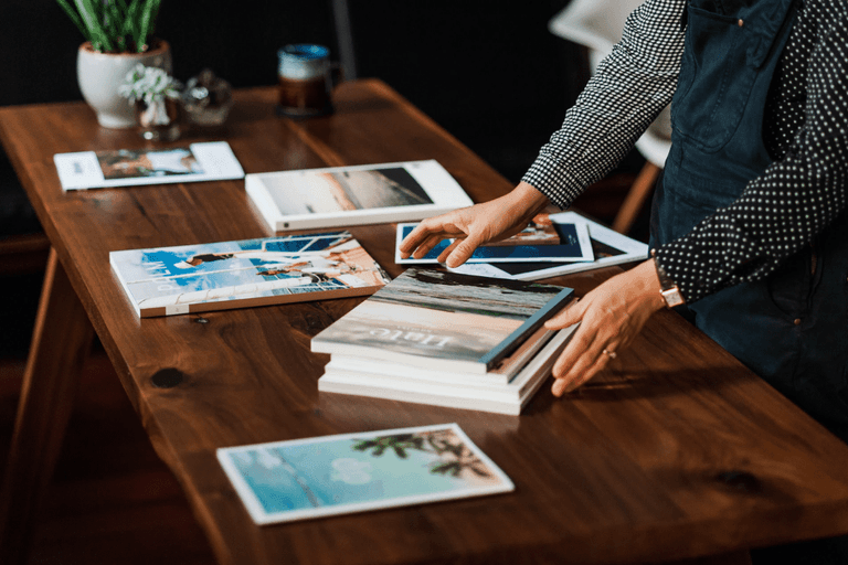 Person arranging coffee table books on a wooden table, with a potted plant and candle in the background.