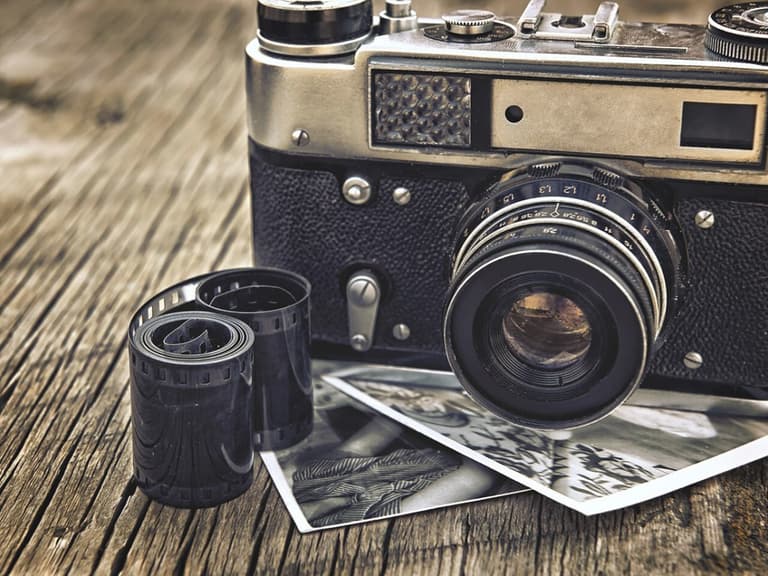 Vintage film camera with black body and silver details on wooden surface, surrounded by film rolls and black-and-white photos.