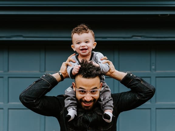 Father with glasses holding smiling baby on shoulders in front of a blue garage door.
