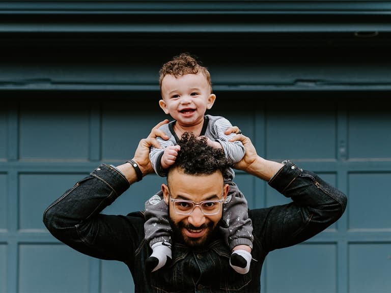 Father with glasses holding smiling baby on shoulders in front of a blue garage door.