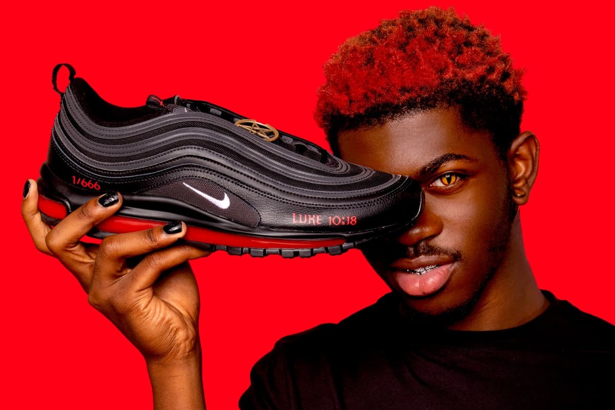 Lil Nas X holds a black and red Nike Air Max 97 with "Luke 10:18" text against a vibrant red background.