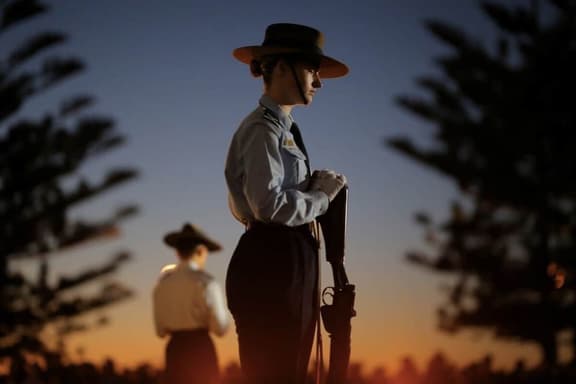 Soldier in uniform holding rifle at sunset, with silhouetted trees and another soldier in the background.