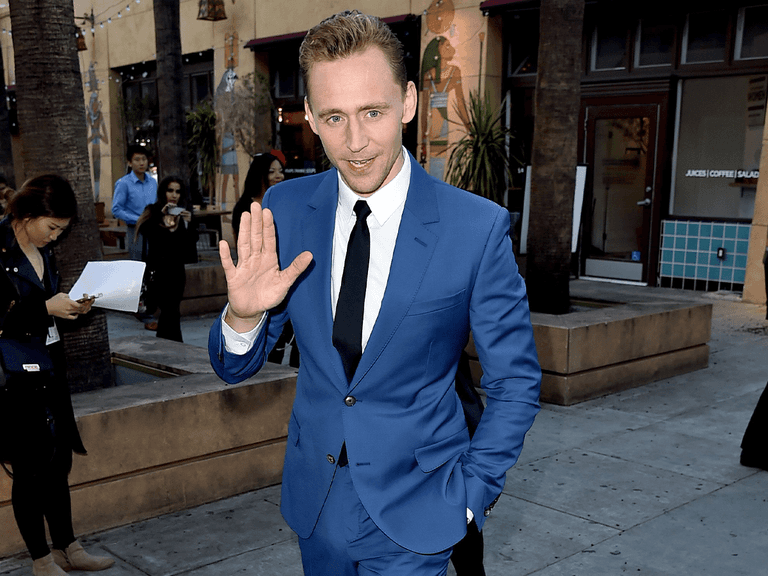 Man in a vibrant blue suit waving, standing outdoors on a city street with palm trees and shops in the background.