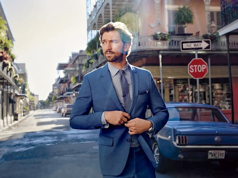 Man in blue suit adjusting jacket on sunlit street with vintage car and historic buildings.