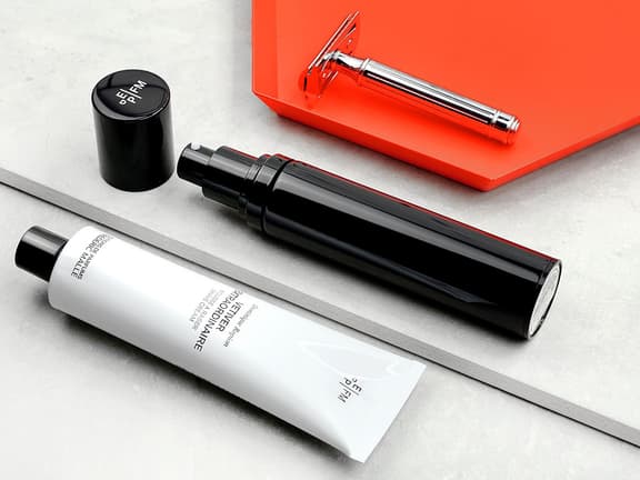 Grooming products including a black pump bottle, white tube, and silver razor on a grey and orange surface.