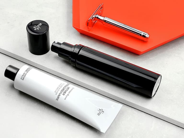 Grooming products including a black pump bottle, white tube, and silver razor on a grey and orange surface.