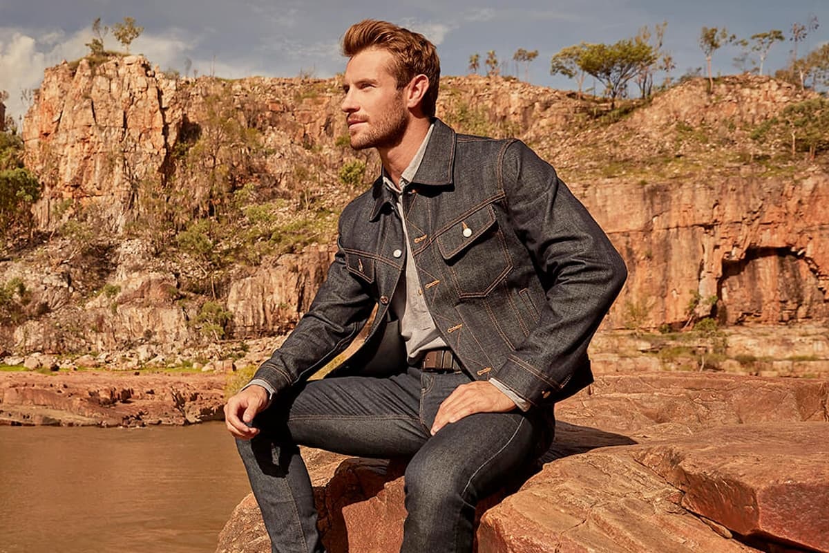 Man in dark denim jacket and jeans sitting on rocky terrain with cliffs and trees in the background.