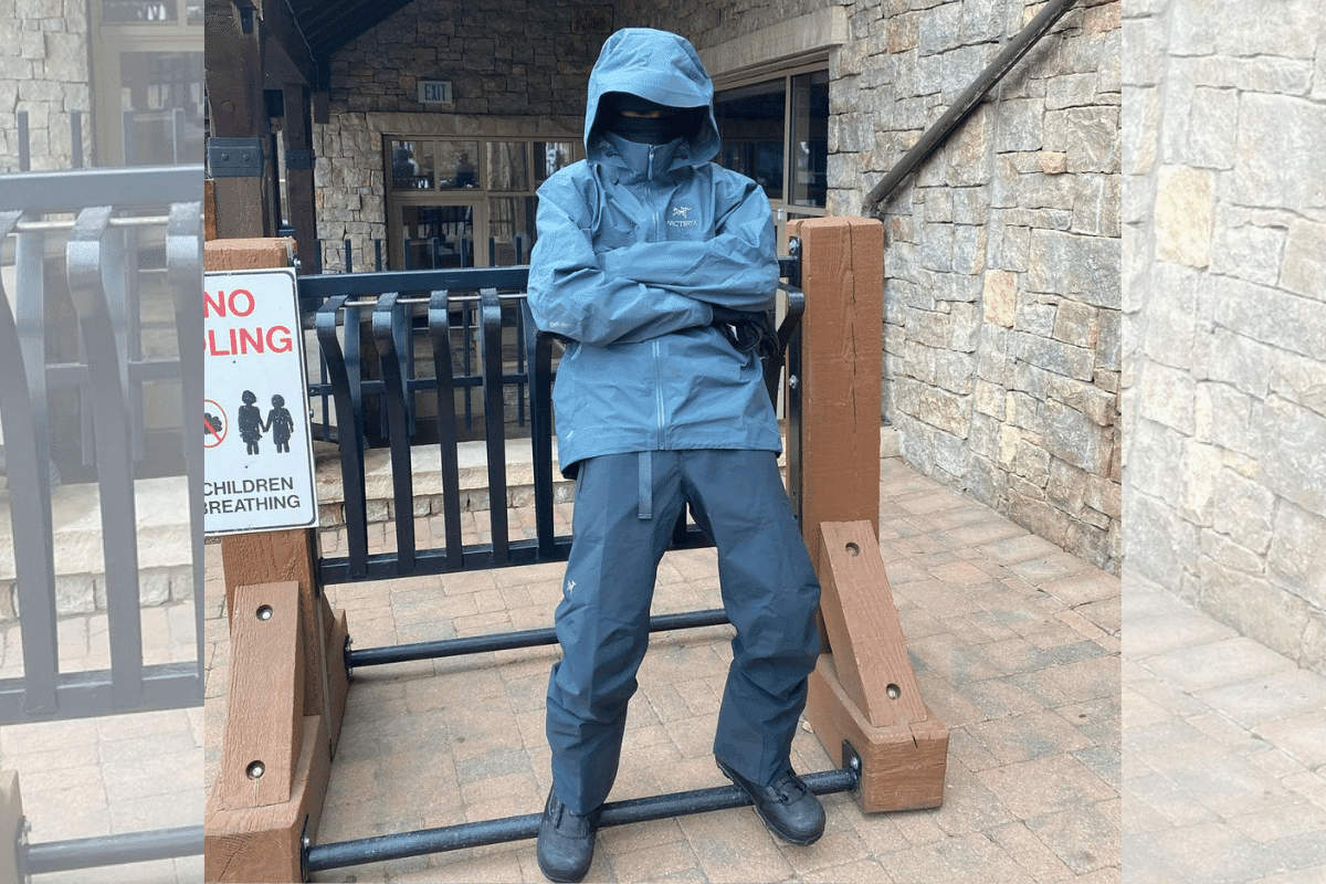 Travis Scott wearing blue Arc'teryx jacket and pants, hood up, leaning against a wooden fence with stone building backdrop.
