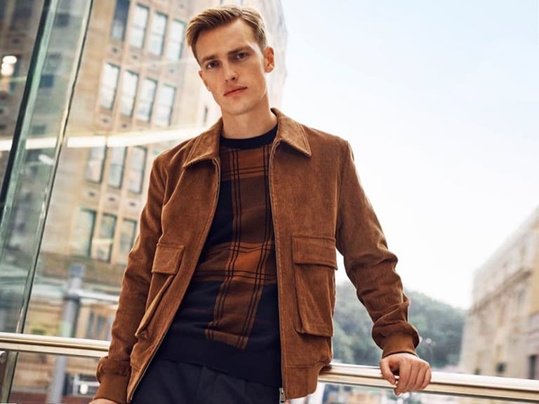 Man wearing a brown suede jacket and plaid shirt stands in a modern urban setting with glass and buildings.