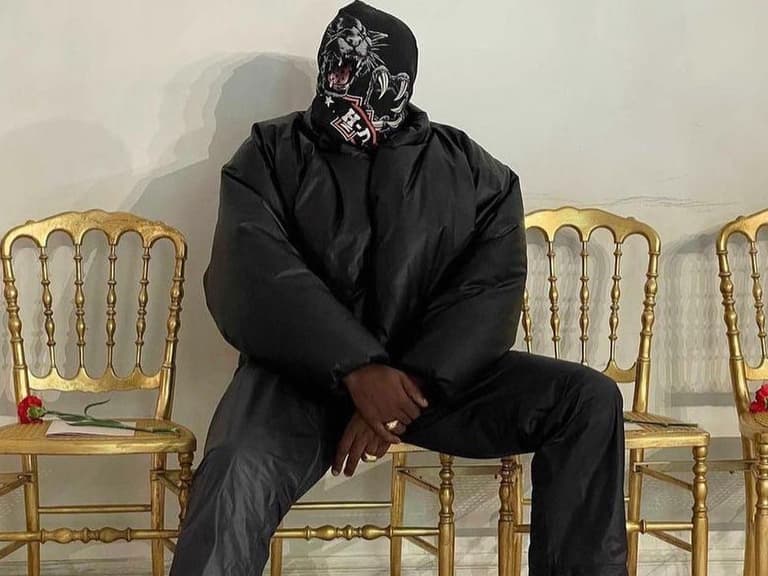 Kanye West wearing a black puffer jacket and mask, sitting on a gold chair with a relaxed pose.