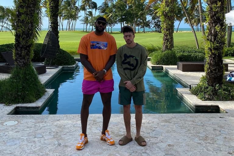 LeBron James in orange shirt and pink shorts stands beside Bad Bunny in green sweatshirt by a pool with palm trees.