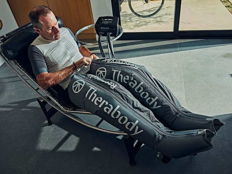Man using Therabody RecoveryAir compression system on legs while seated in a modern chair, with natural light streaming in.