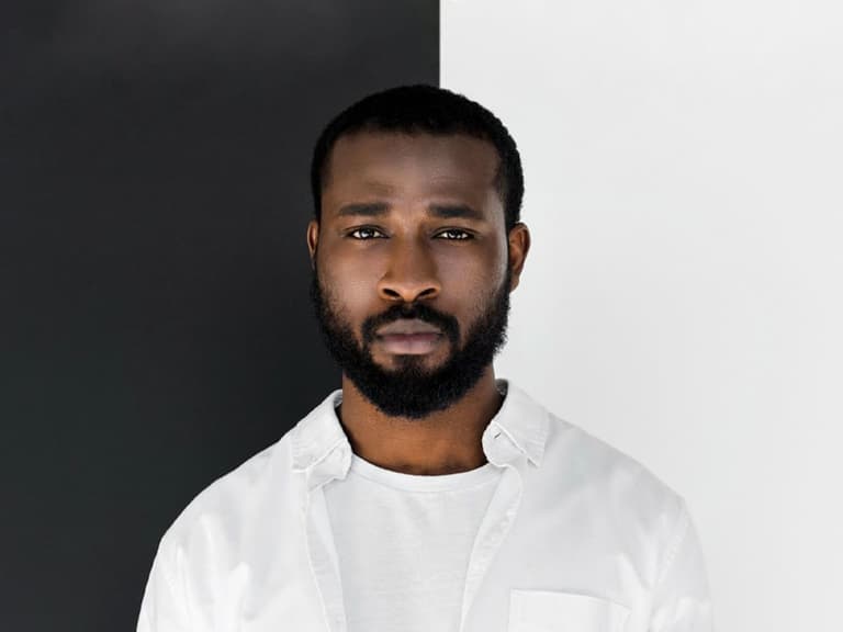 Man in white shirt standing against a black and white split background, looking directly at the camera.