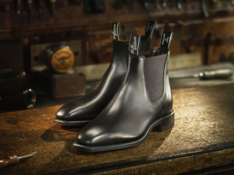 R.M. Williams black leather work boots on a rustic wooden table in a workshop setting.