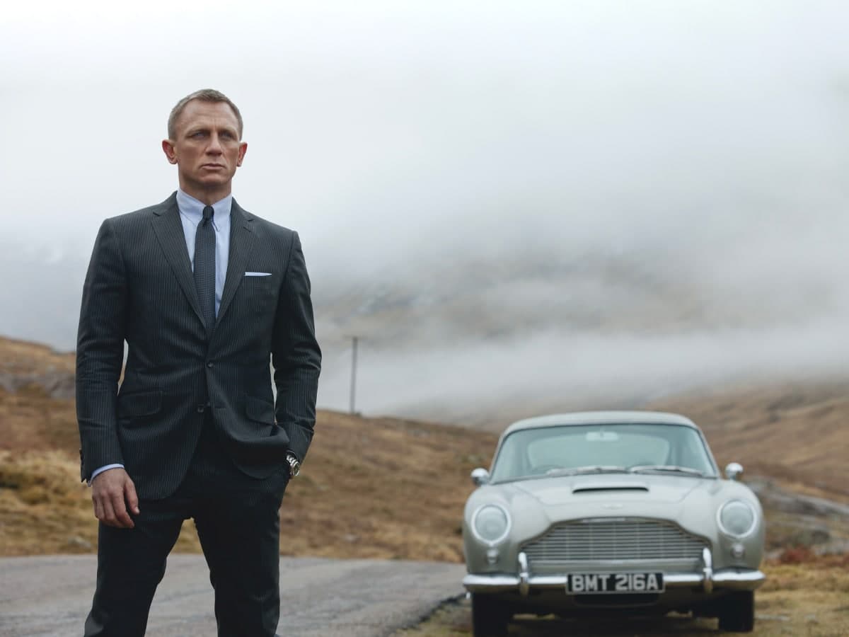 Daniel Craig stands in front of a classic silver Aston Martin DB5 on a foggy rural road.